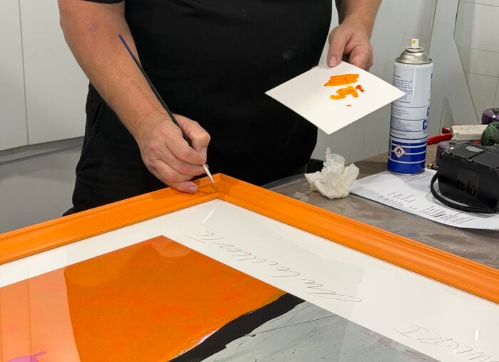 Man holding paintbrush painting the corner of a custom orange frame in the Dawsons Framing workshop Ashmore
