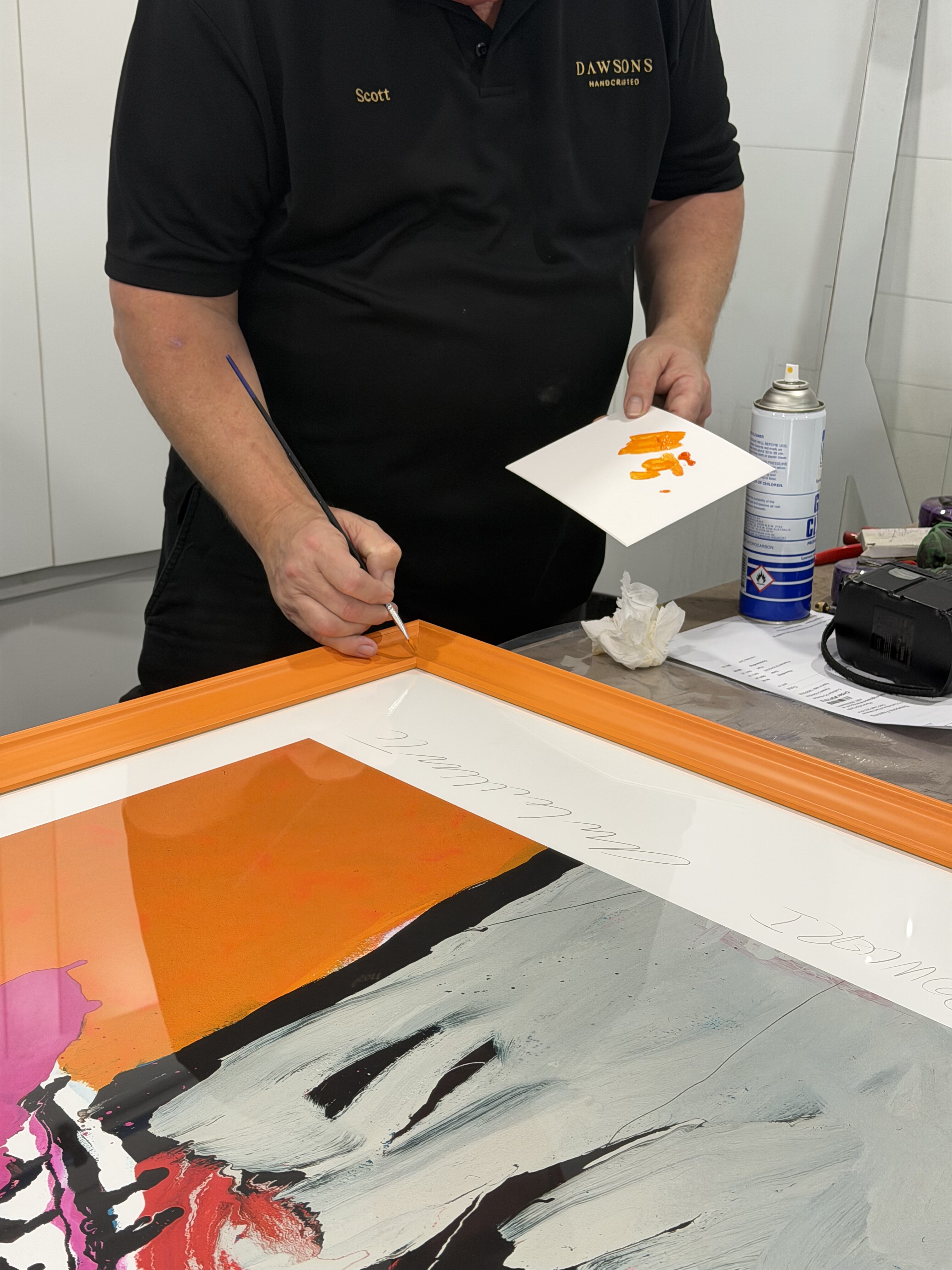 Man holding paintbrush painting the corner of a custom orange frame in the Dawsons Framing workshop Ashmore